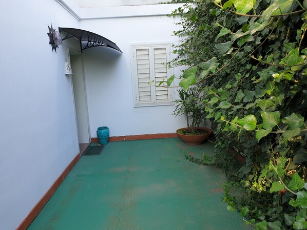 Un pequeño patio interior con suelo verde, una planta en maceta, un paraguas decorativo y una pared blanca con una ventana.