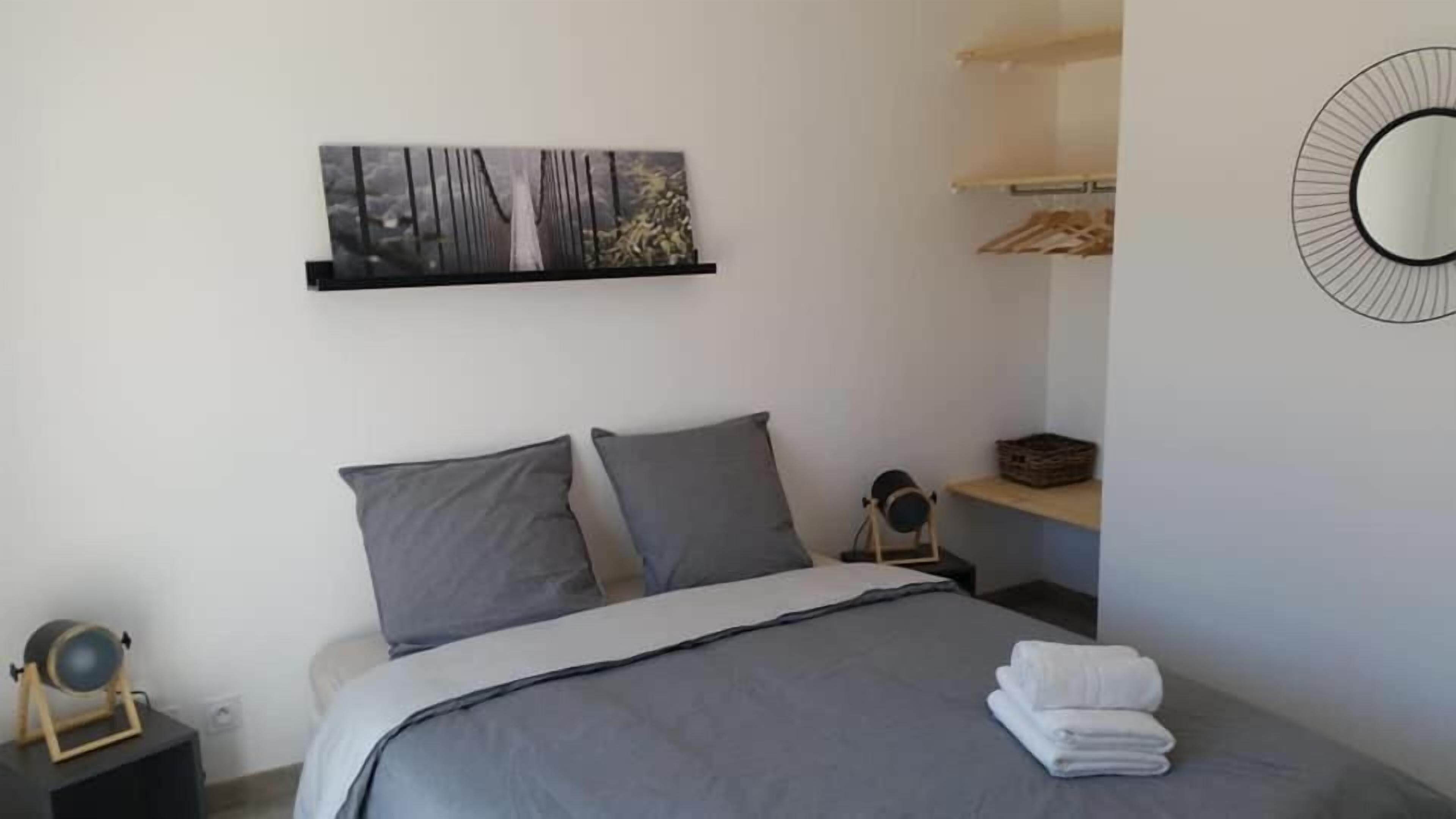 Un dormitorio con cama, dos almohadas, una mesita de noche con lámpara, un estante en la pared con ropa y un cuadro.