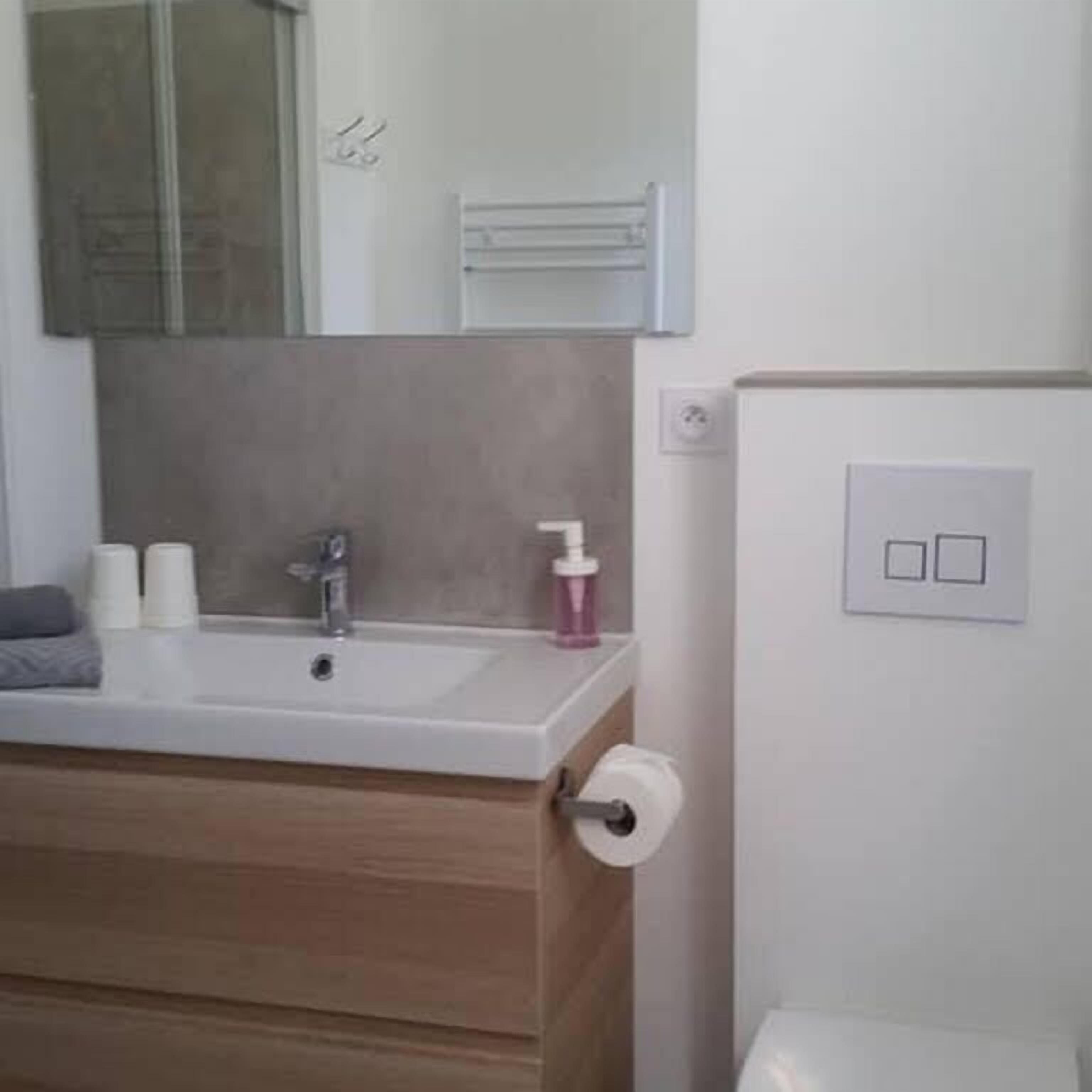 A modern bathroom with a wooden vanity, a white sink, and a mirror above it. There are toiletries on the counter and a towel rack above the mirror.