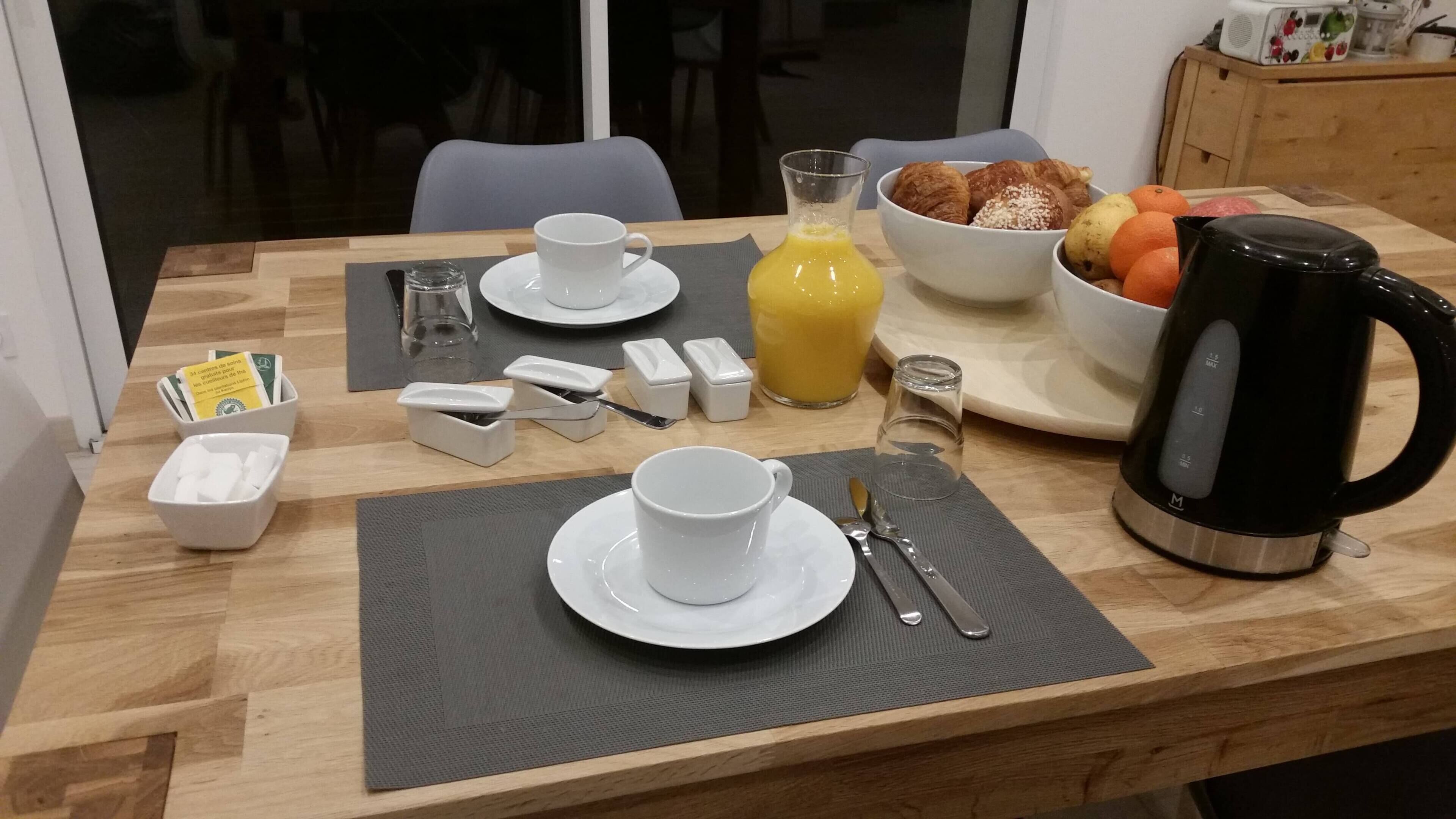 Ein Frühstückstisch gedeckt mit Tasse, Untertasse, Glas, Saft, Croissants, Orangen und einer Kaffeemaschine.