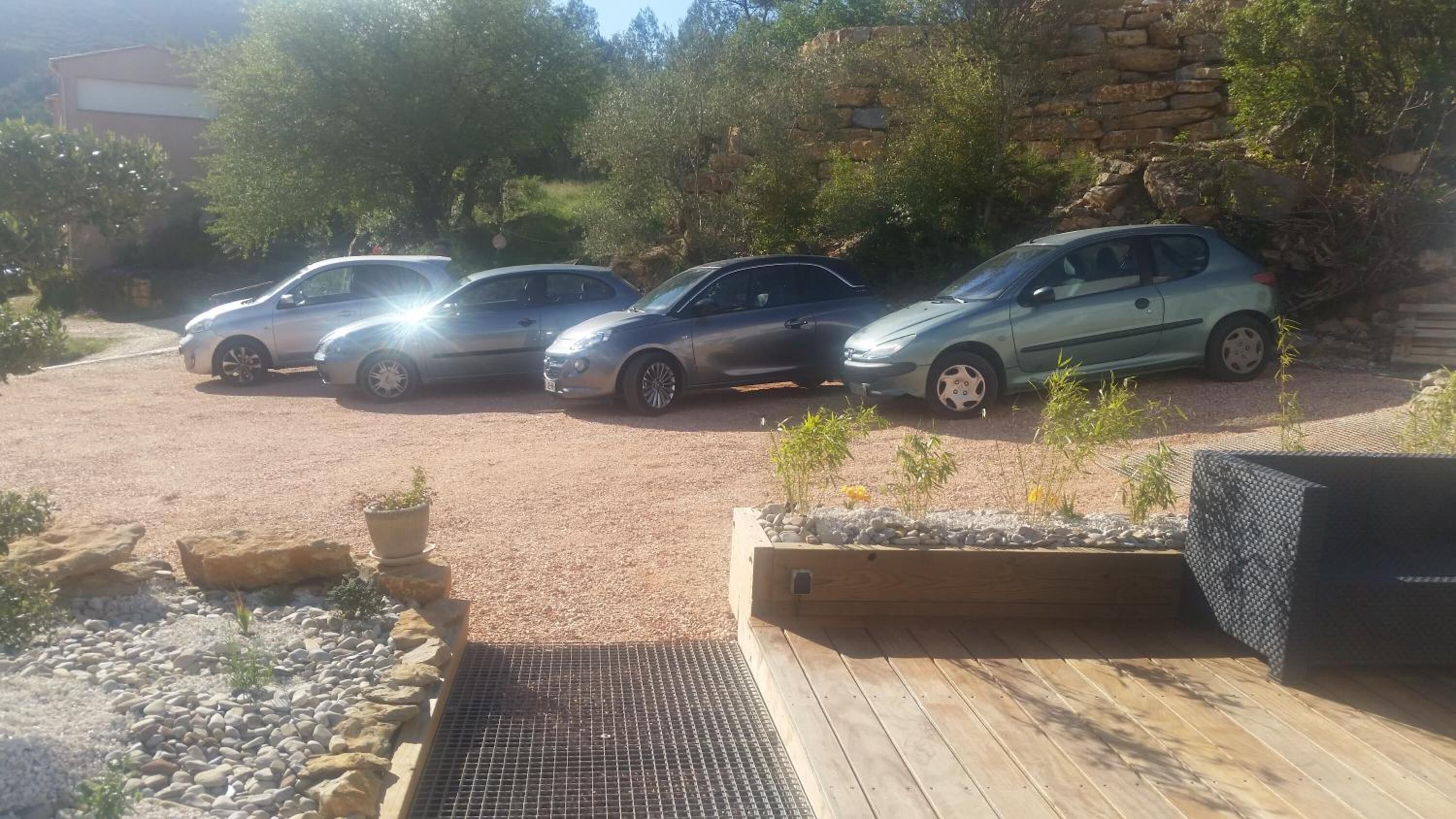 Un área de estacionamiento con cuatro coches aparcados en fila, una terraza de madera y un muro de piedra al fondo.