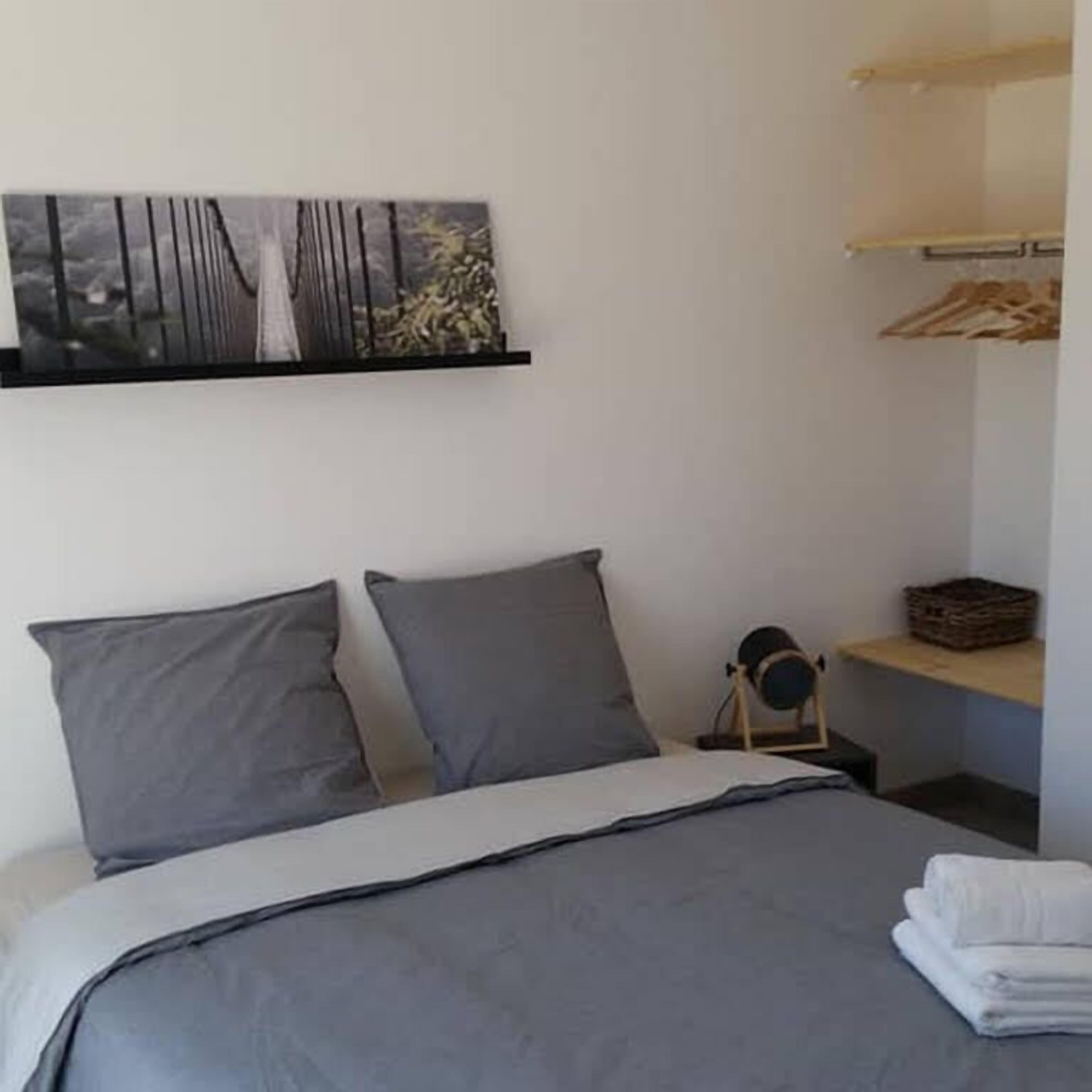 Un dormitorio con una cama, dos almohadas, una mesita de noche con una lámpara y un estante en la pared con una cesta.