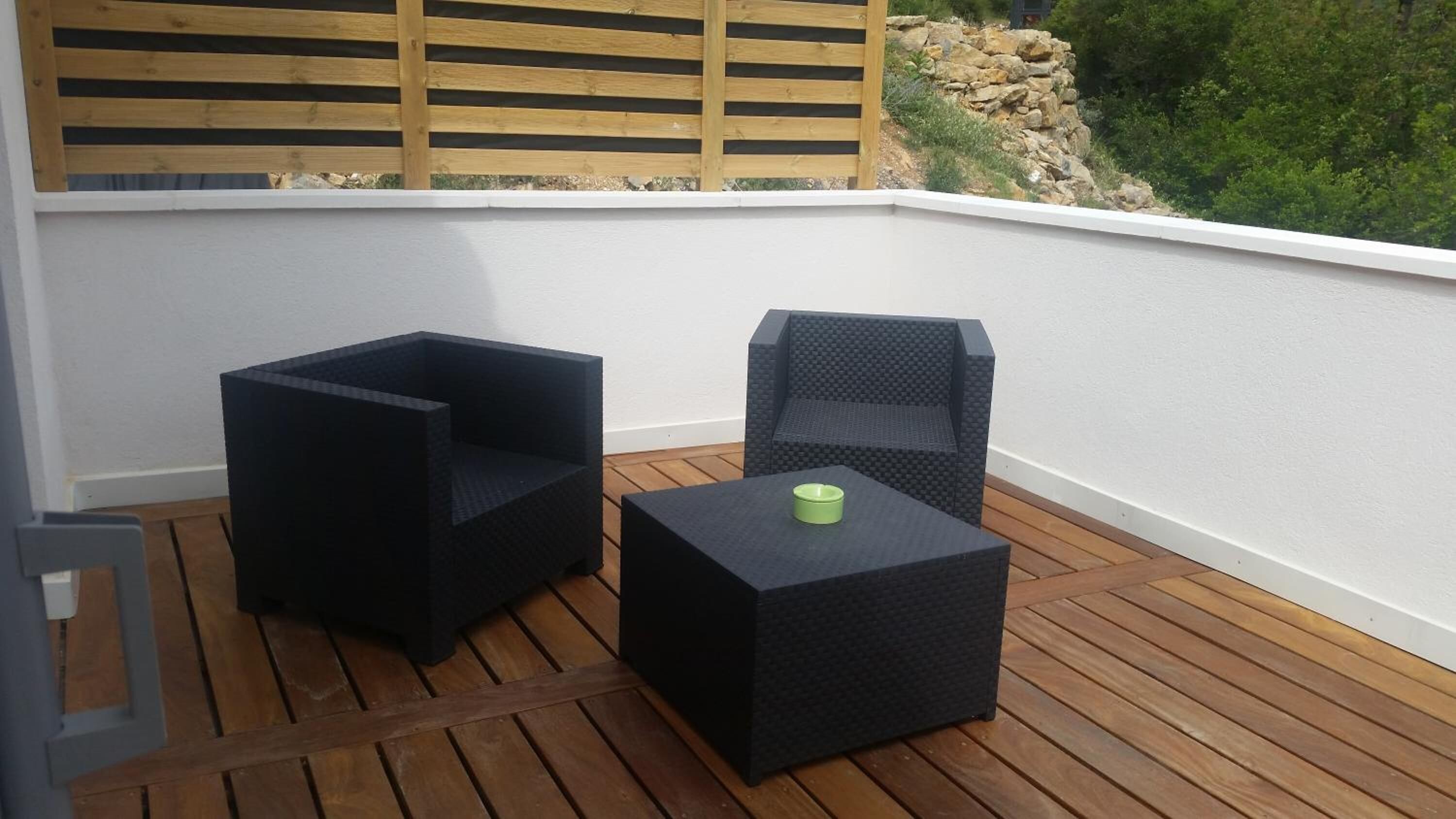A balcony with two black square chairs and a small table, a wooden fence, and a view of greenery.
