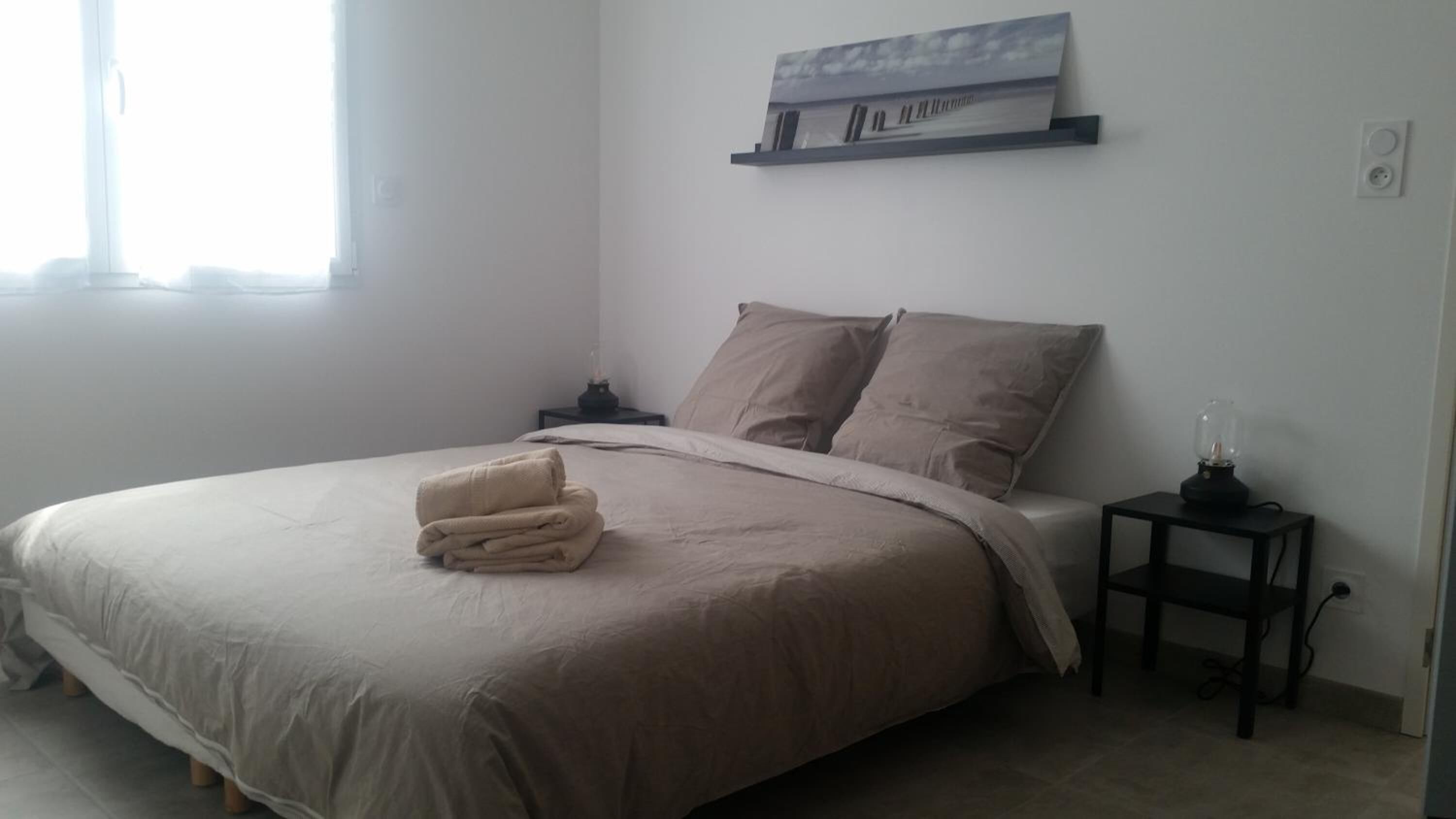 A bedroom with a large bed, two pillows, a bedside table with a lamp, and a wall-mounted shelf with a picture.