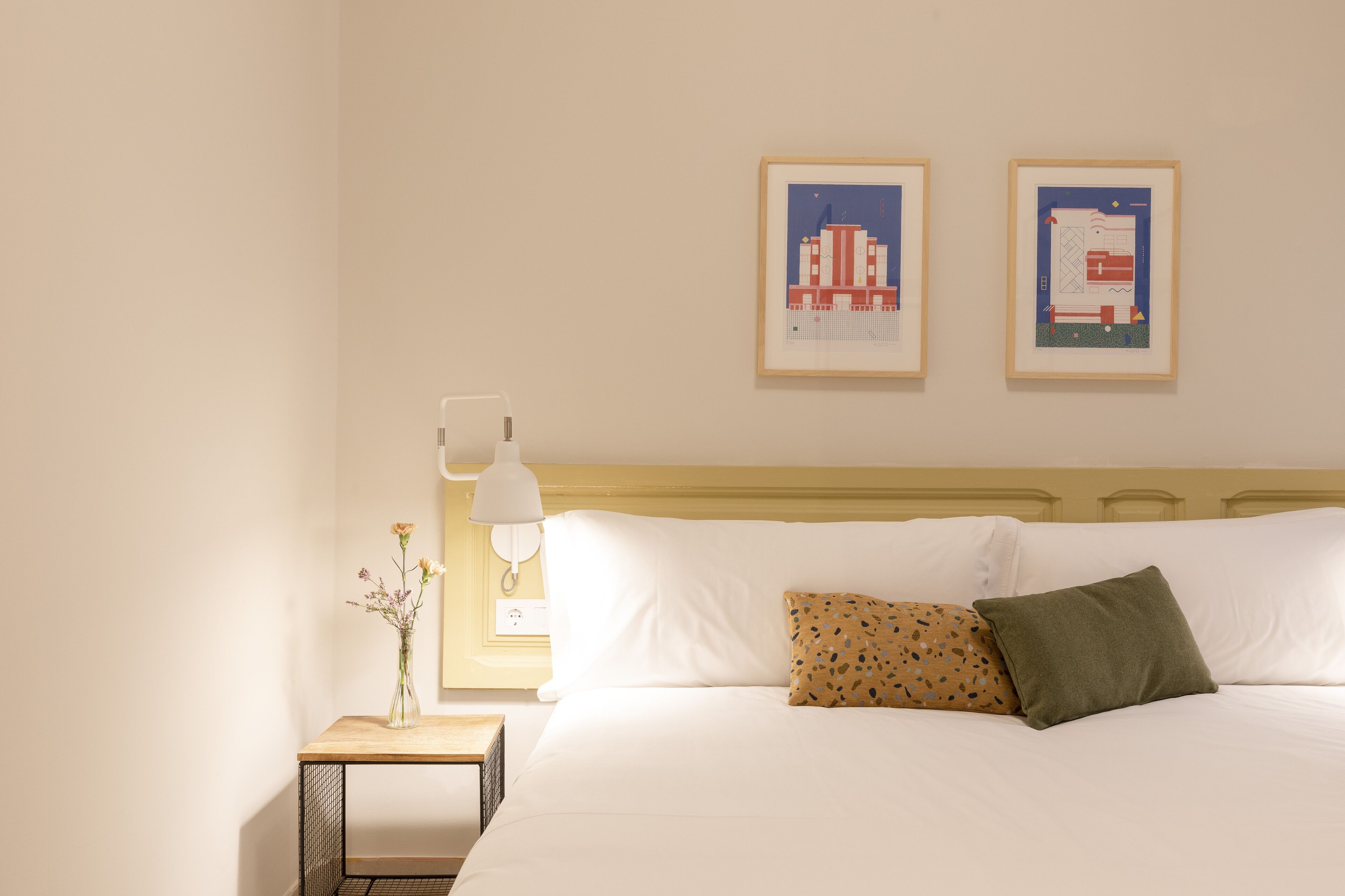 A neatly made bed with white linens and two pillows, a small bedside table with a vase of flowers, and two framed artworks on the wall.