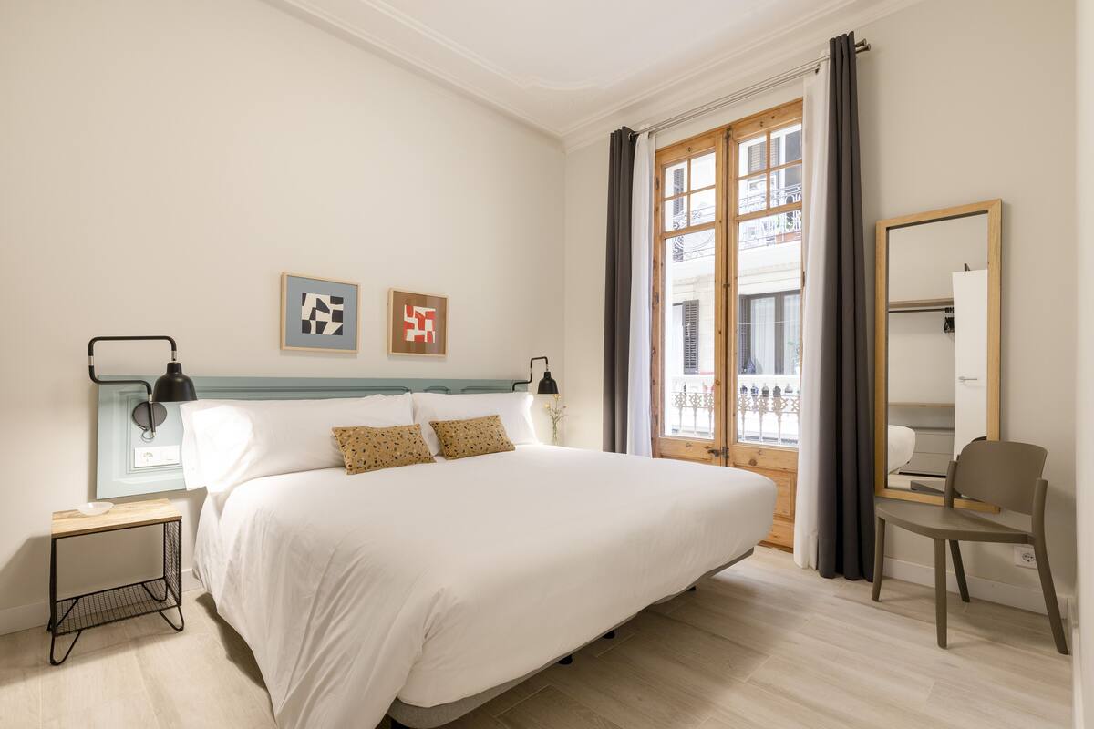 Une chambre à coucher avec un grand lit, des tables de chevet, un miroir et une fenêtre avec des rideaux.