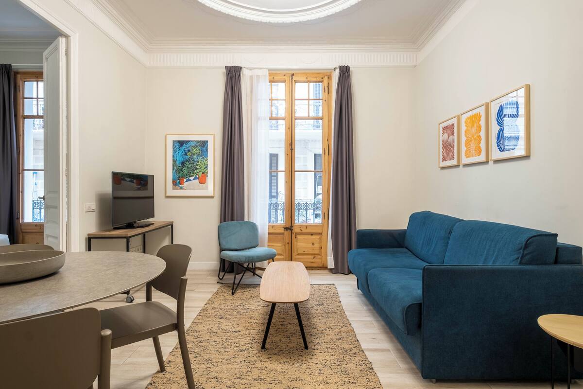 Un salon avec un canapé bleu, une table à manger avec des chaises, un téléviseur posé sur un meuble et une grande fenêtre avec des rideaux.