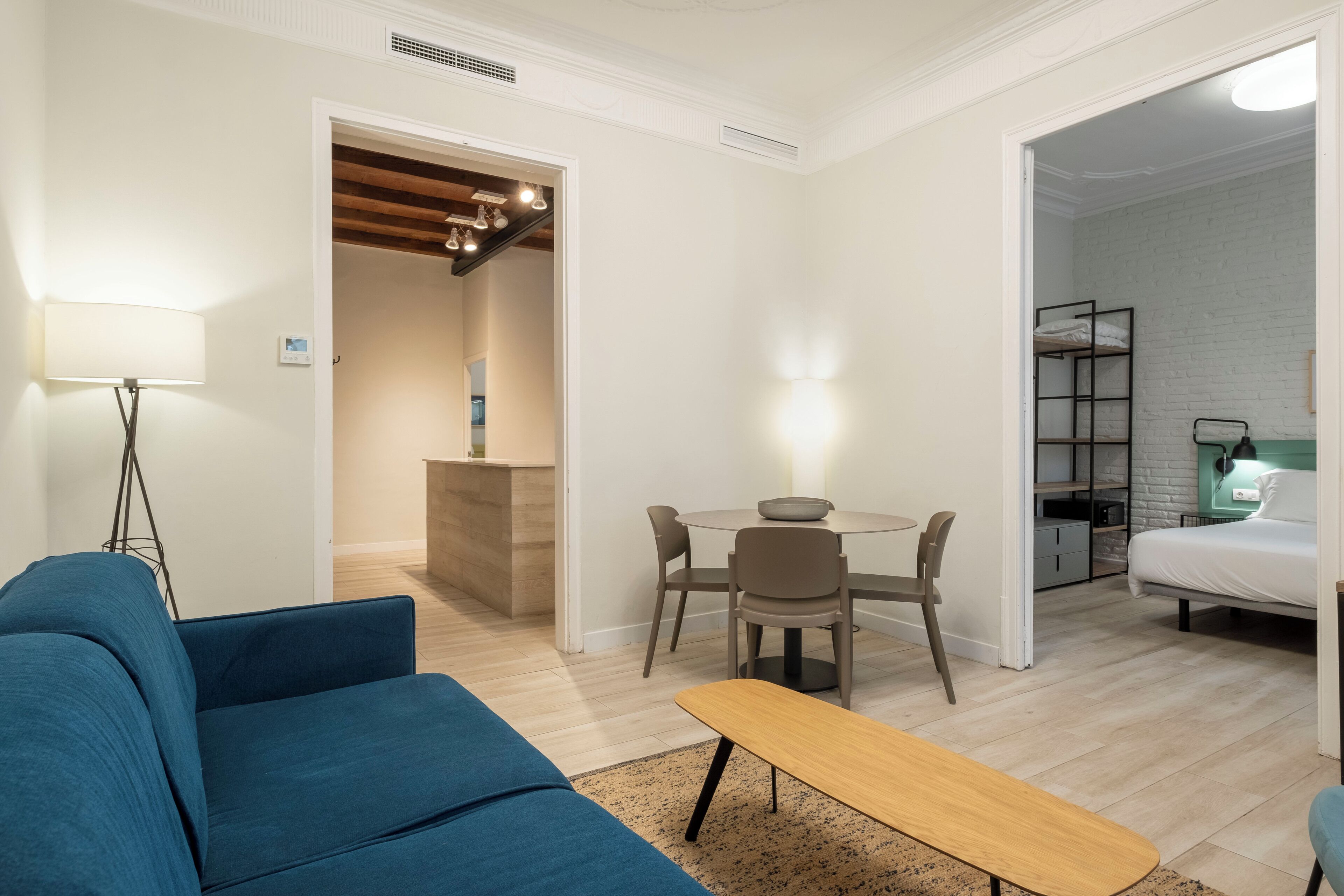 A modern living room with a blue sofa, a wooden bench, a dining table with chairs, and a small kitchen area with a bench.