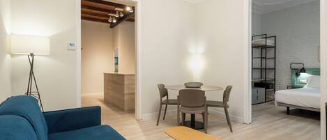 A modern living room with a blue sofa, a wooden bench, a dining table with chairs, and a small kitchen area with a bench.