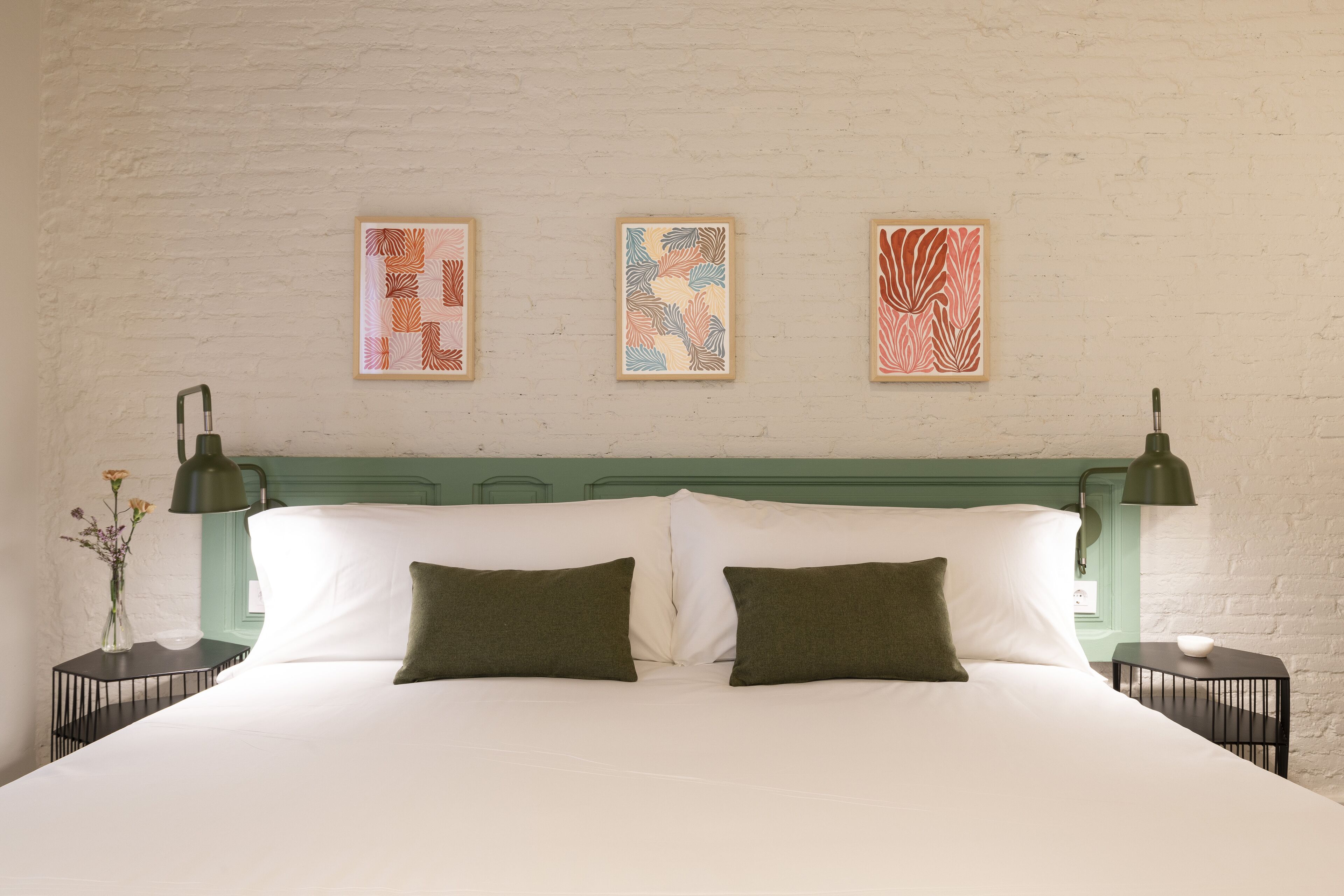Ein Bett mit weißer Bettwäsche und zwei grünen Kissen, seitlich flankiert von schwarzen Nachttischen mit Lampen. Darüber an der Wand hängen drei gerahmte Kunstwerke.