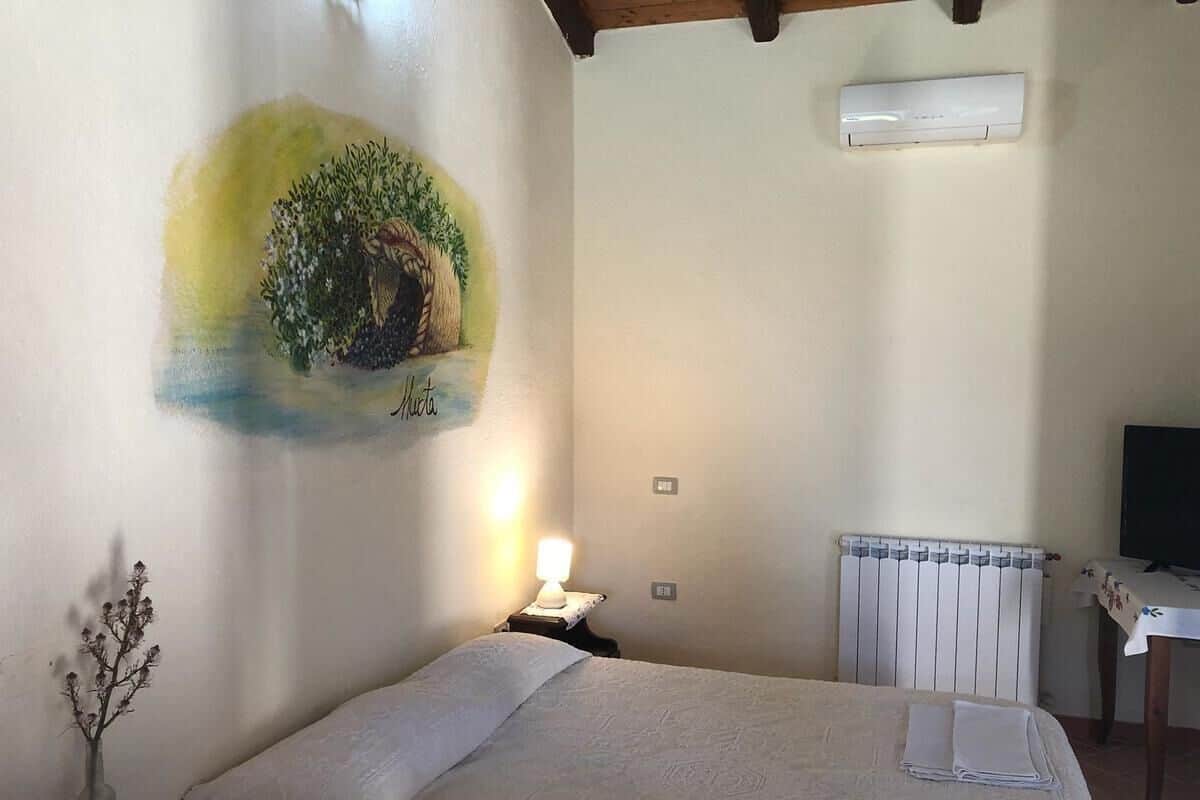 A bedroom with a bed, a nightstand with a lamp, a radiator, a TV, and a wall painting.