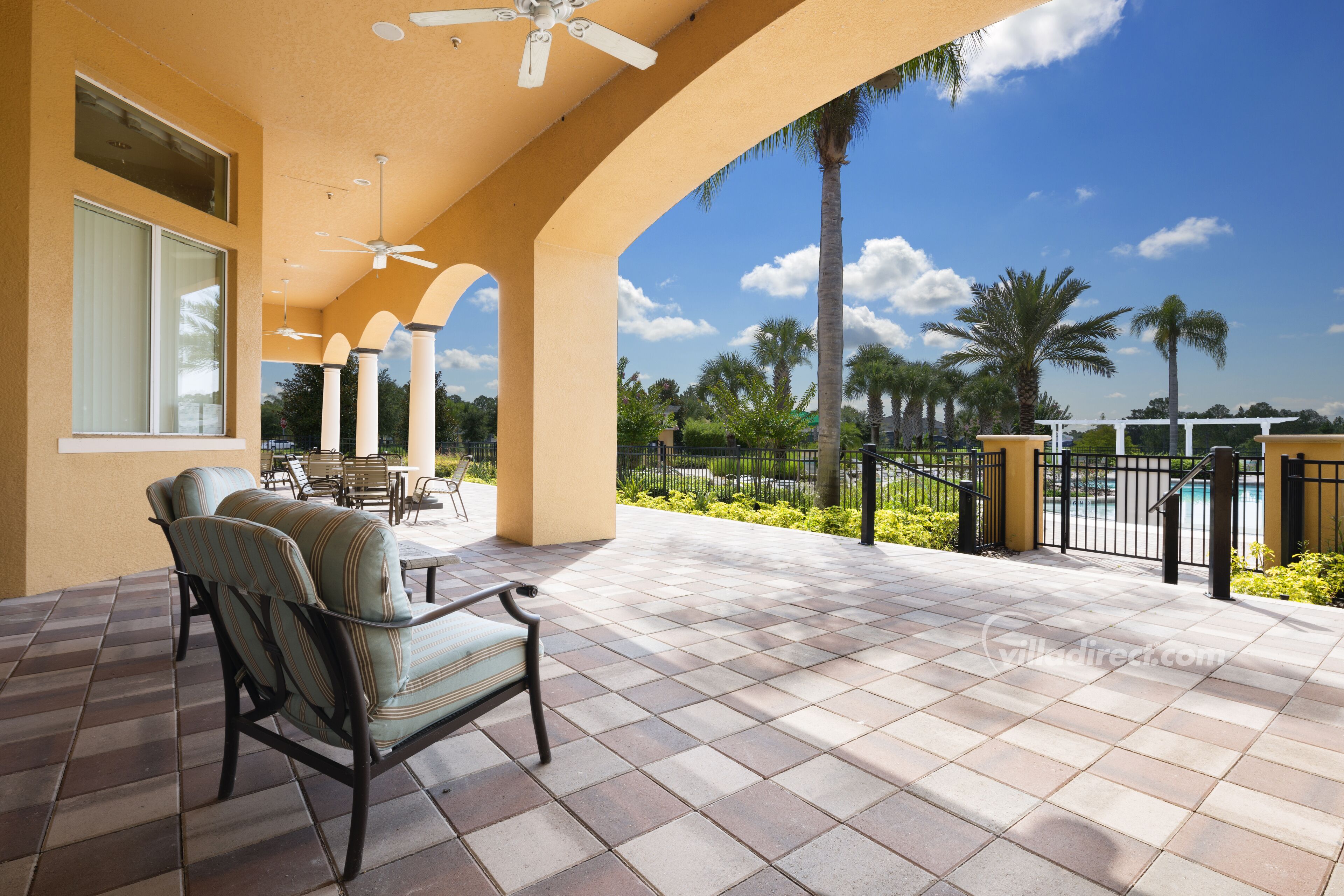 A covered patio with a seating area, a ceiling fan, and a view of a pool and palm trees.