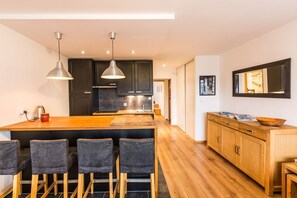 A modern kitchen with dark cabinets, a wooden island, bar stools, pendant lights, and a wooden cabinet with a bowl on top.