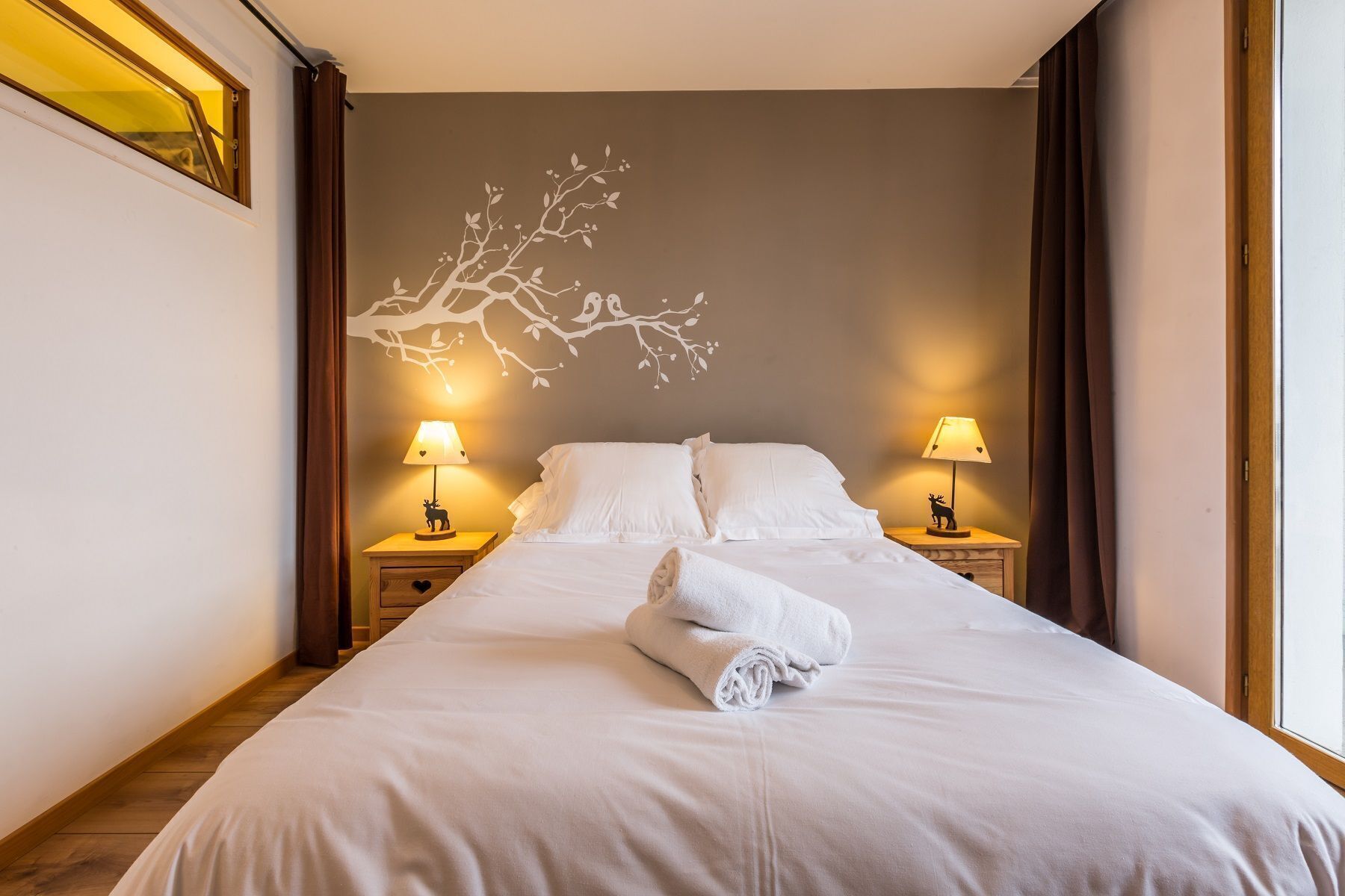 A neatly made bed with white linens and a rolled towel, two bedside lamps, and a wall mural of a tree branch.