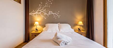 A neatly made bed with white linens and a rolled towel, two bedside lamps, and a wall mural of a tree branch.