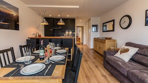 Eine moderne Küche mit dunklen Schränken und Pendelleuchten, ein Essbereich mit einem Holztisch, gedeckt für vier Personen, und ein Wohnzimmer mit einer Couch und einer Wanduhr.