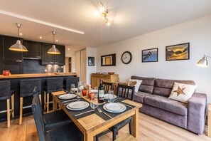 A modern kitchen with dark cabinets and pendant lighting, a dining area with a wooden table set for four, and a living room with a purple sofa and wall art.