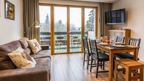 Ein gemütliches Wohnzimmer mit Holzboden, einer braunen Couch, einem Esstisch mit Stühlen und einem an der Wand befestigten Fernseher.