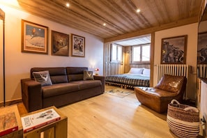 Ein gemütliches Zimmer mit Holzbalkendecke, einem braunen Ledersofa, einem Bett und einem Sessel. An den Wänden hängen Bilder in Rahmen und auf dem Boden steht ein Korb.