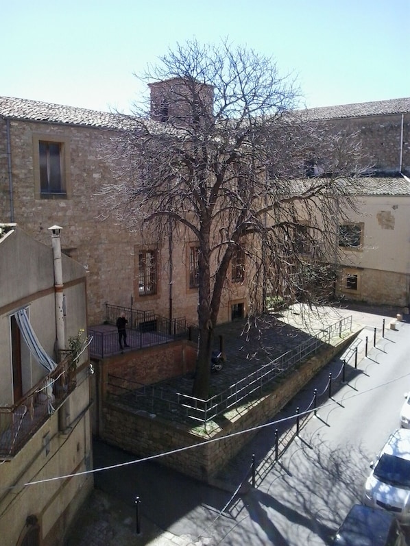 A tree-lined courtyard with stone buildings, a paved area, and a metal fence. - La Dea Bendata (Piazza Armerina)