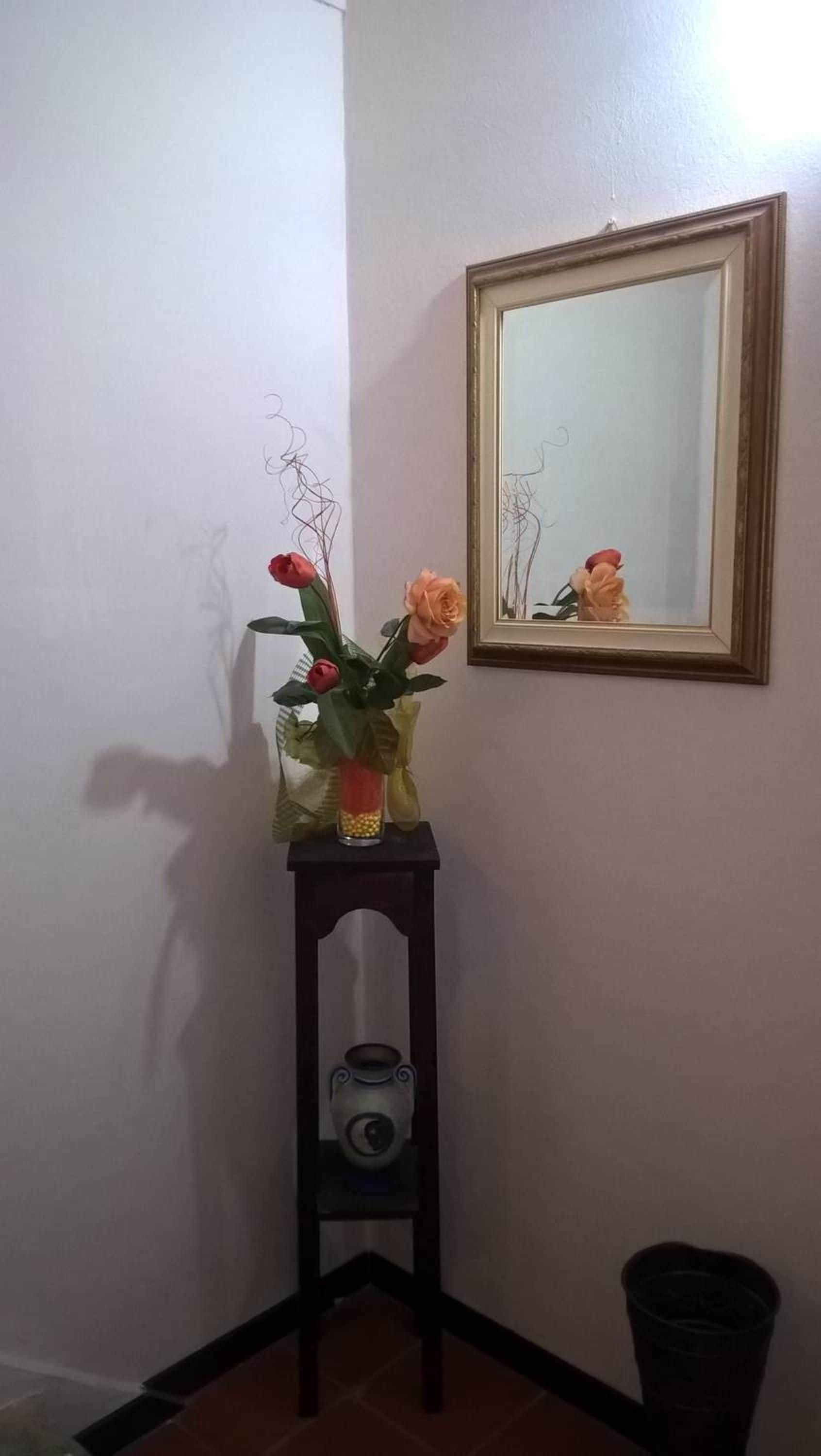Un coin avec une petite table sur laquelle est posé un vase de fleurs, un miroir encadré accroché au mur et une poubelle.