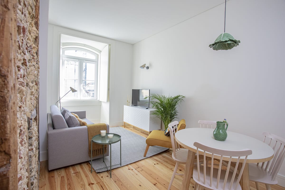 A modern living room with a sofa, a dining table, and chairs. There is a television on a white cabinet, a plant, and a green hanging light fixture.