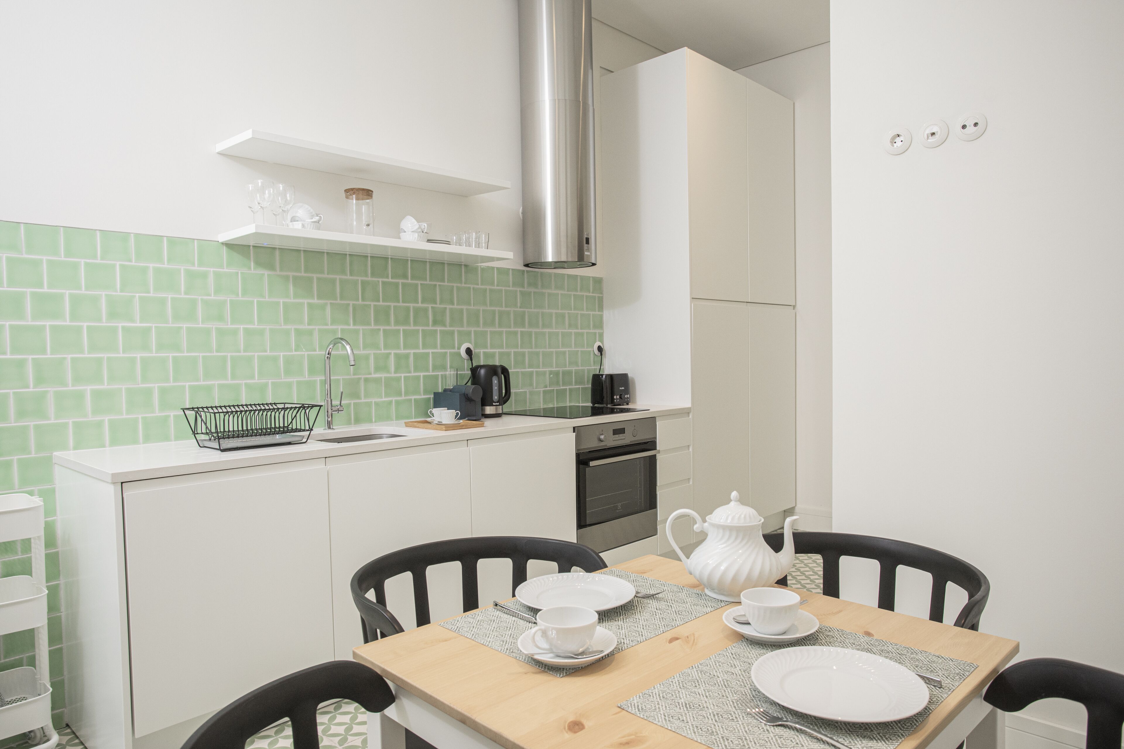 Een compacte keuken met witte kasten, een groene betegelde spatwand, een roestvrijstalen afzuigkap en een eethoek met een houten tafel en zwarte stoelen.