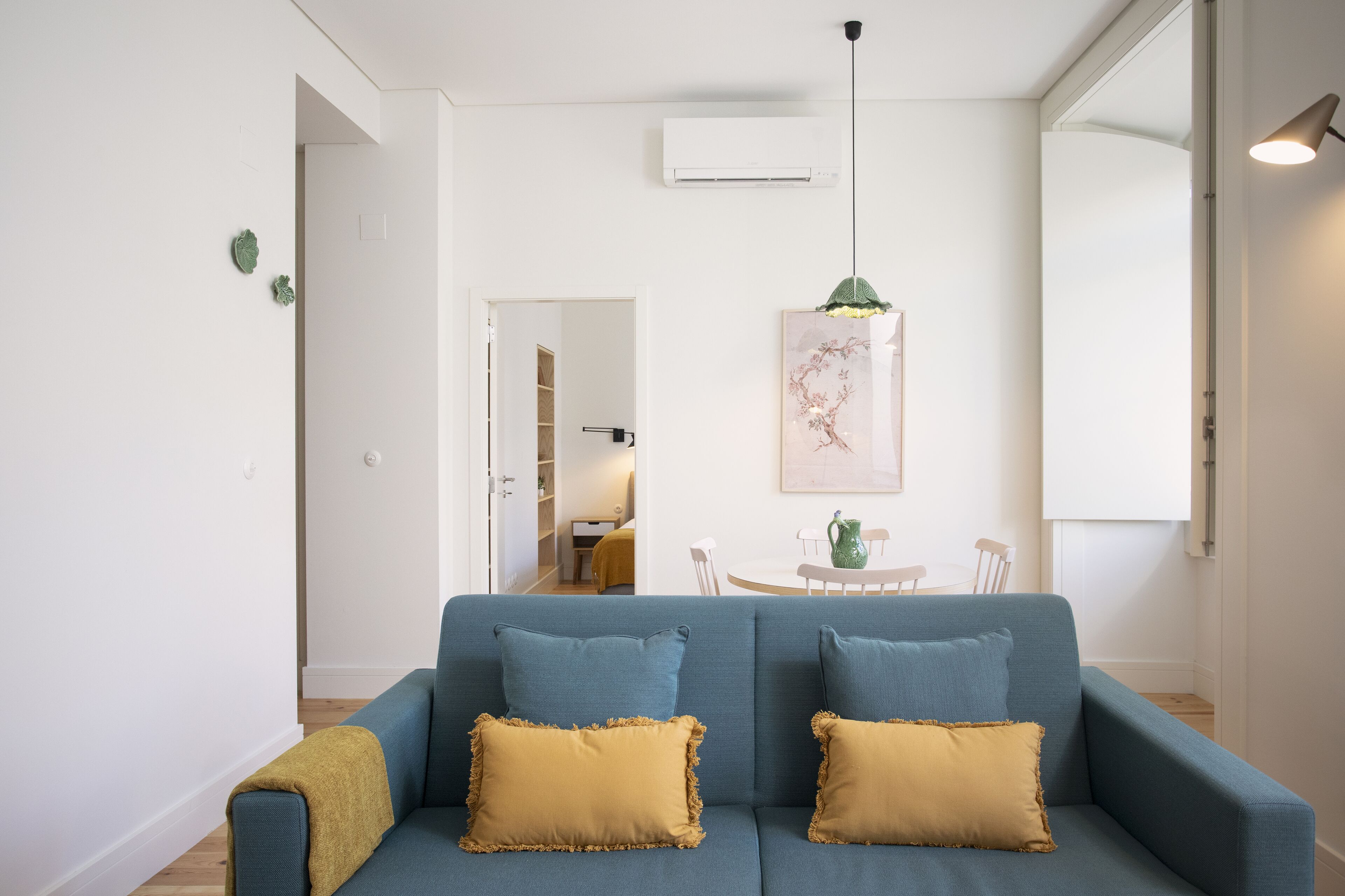 Ein modernes Wohnzimmer mit einem blauen Sofa, zwei senfgelben Kissen, einer grünen Pendelleuchte und einem Essbereich im Hintergrund.