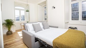A modern bedroom with a bed, a grey sofa, a small table, a potted plant, and a window with a view of buildings. - Montebelo Lisbon Downtown Apartments (Lisbon)