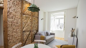 A living room with a stone wall, a grey sofa, a yellow armchair, a white table with a vase, and a hanging light fixture. - Montebelo Lisbon Downtown Apartments (Lisbon)