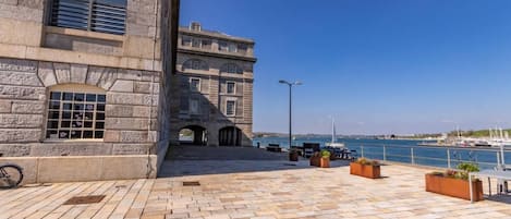 Un edificio frente al agua con arquitectura de piedra, una terraza empedrada y vistas al mar.