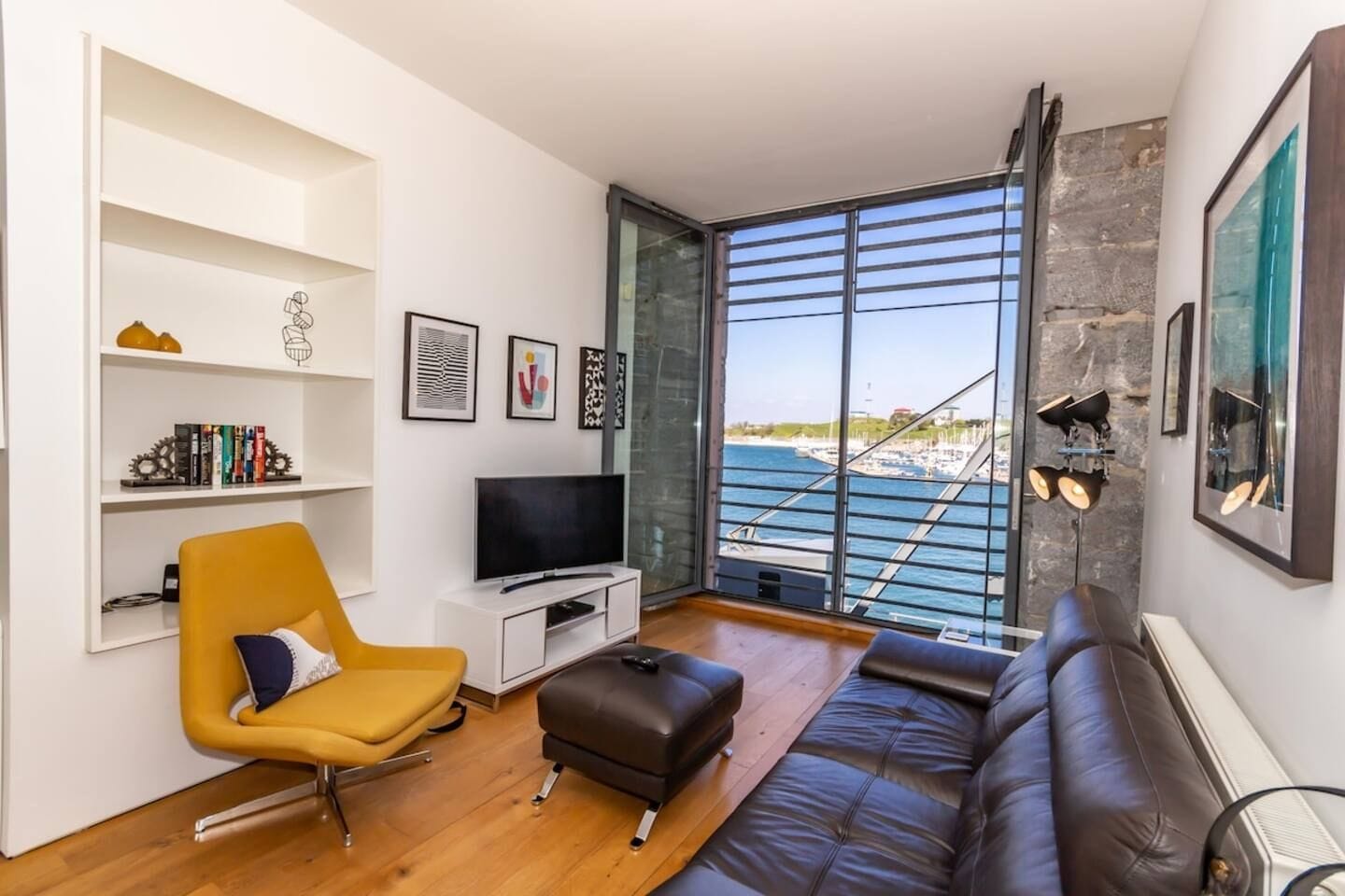 A modern living room with a television, a yellow chair, a brown sofa, and a large window overlooking a marina.