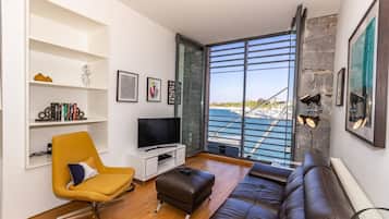 A modern living room with a television, a yellow chair, a brown sofa, and a large window overlooking a marina.