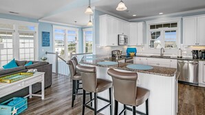A modern kitchen with white cabinets, a granite countertop, and stainless steel appliances. There is a bar area with stools and a view of the outside. - "A Penny From Heaven" 2nd row luxury 4 bedroom with pool (Holden Beach)