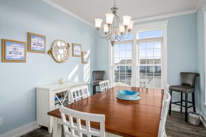 Un coin repas avec une table en bois, des chaises blanches, un lustre et des œuvres d’art encadrées accrochées au mur.