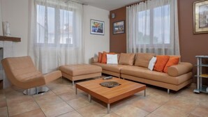 Una sala de estar moderna con un sofá, un sillón, un reposapiés, una mesa de centro y ventanas amplias con cortinas ligeras.