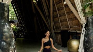 A person meditating in a thatched-roof structure with a mat, surrounded by large vases and a yoga mat visible in the background.