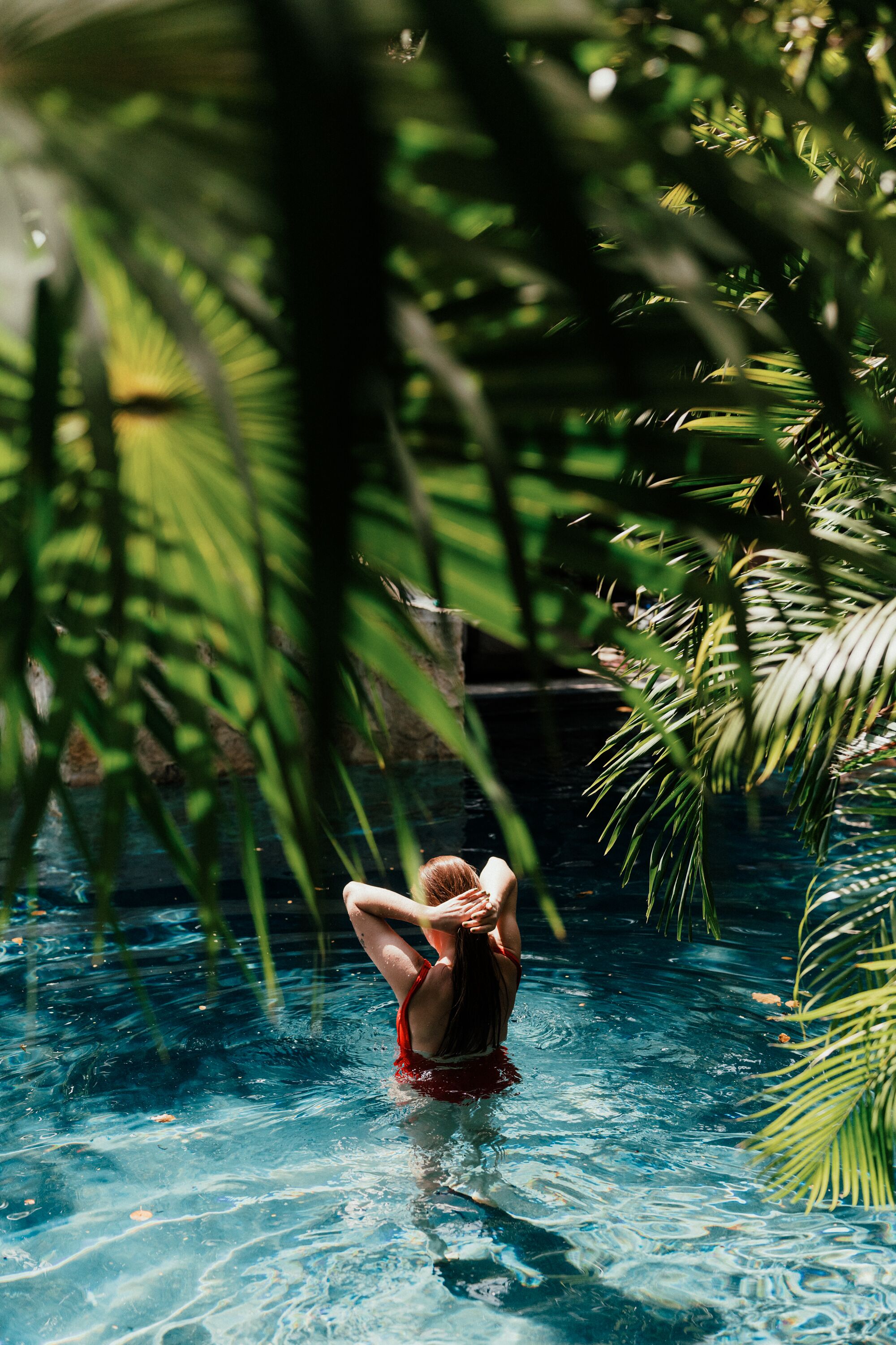 A person in a red swimsuit standing in a clear pool surrounded by lush green foliage.