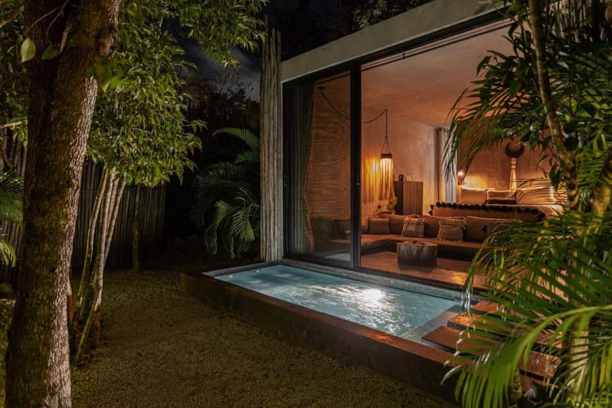 Una casa moderna con un ventanal de vidrio que da a una sala de estar, una pequeña piscina exterior y abundante vegetación.