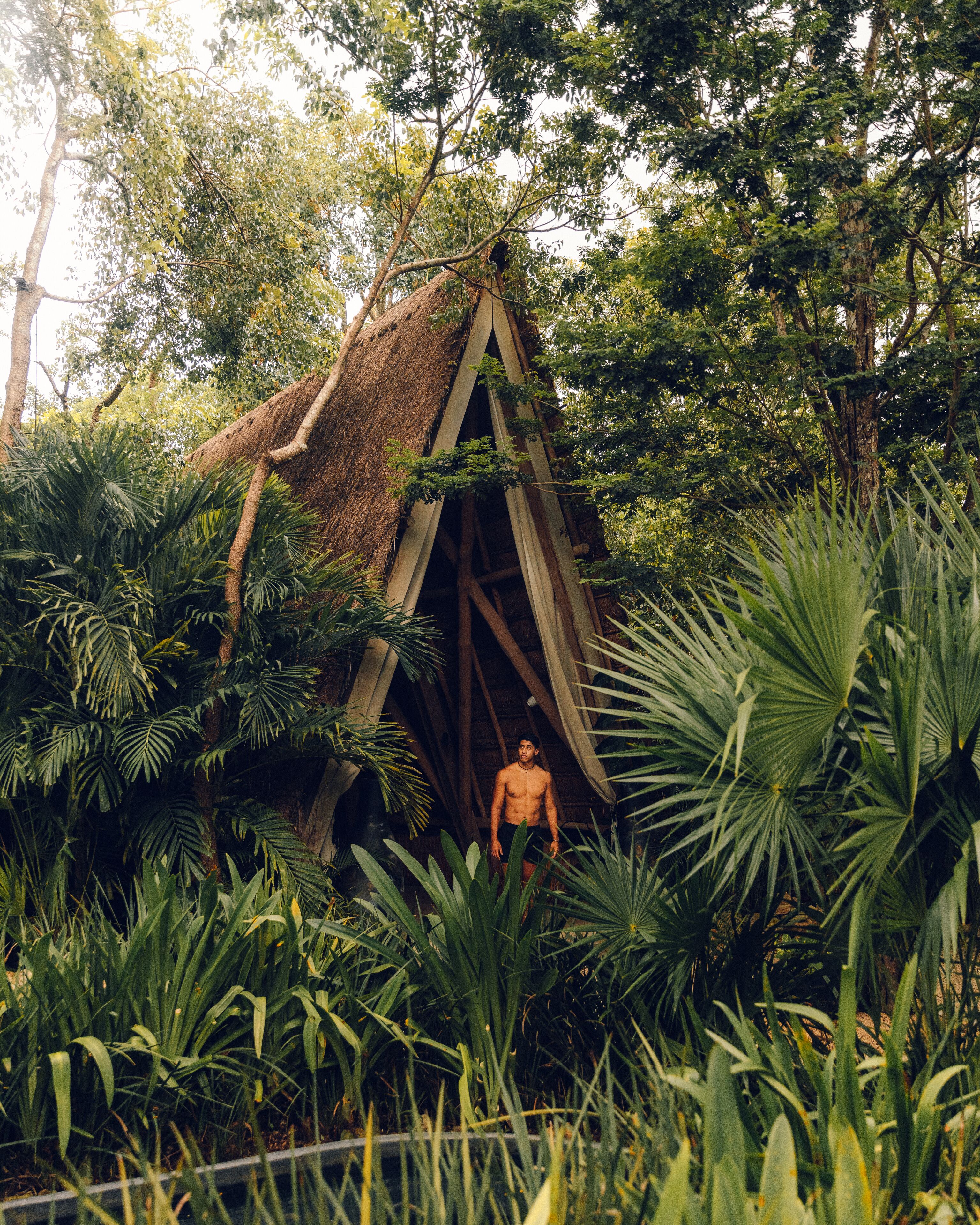 Um homem em um ambiente natural com uma construção de colmo rodeada por vegetação exuberante.