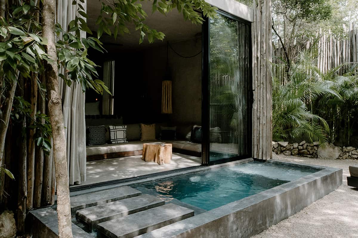 Un área de piscina moderna al aire libre con cerramiento de vidrio, escalones de piedra y una zona de estar con cojines.