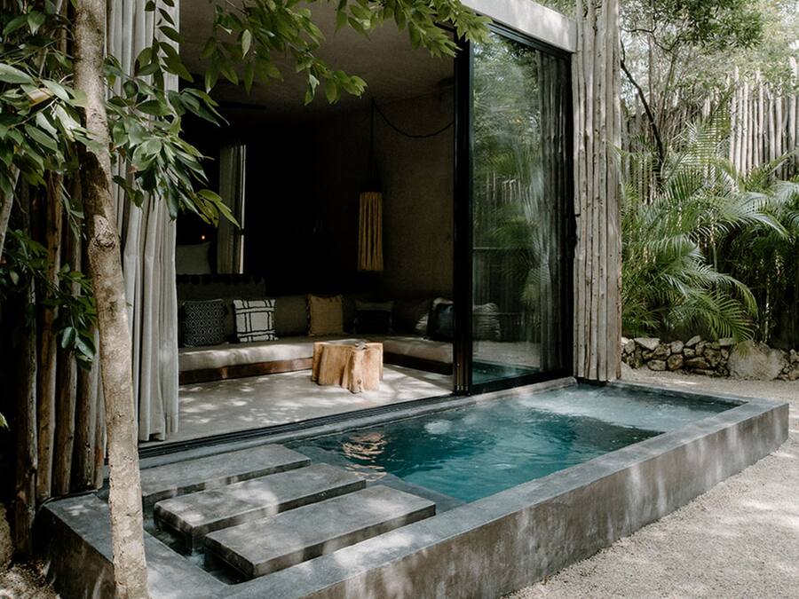 Un área de piscina moderna al aire libre con cerramiento de vidrio, escalones de piedra y una zona de estar con cojines.