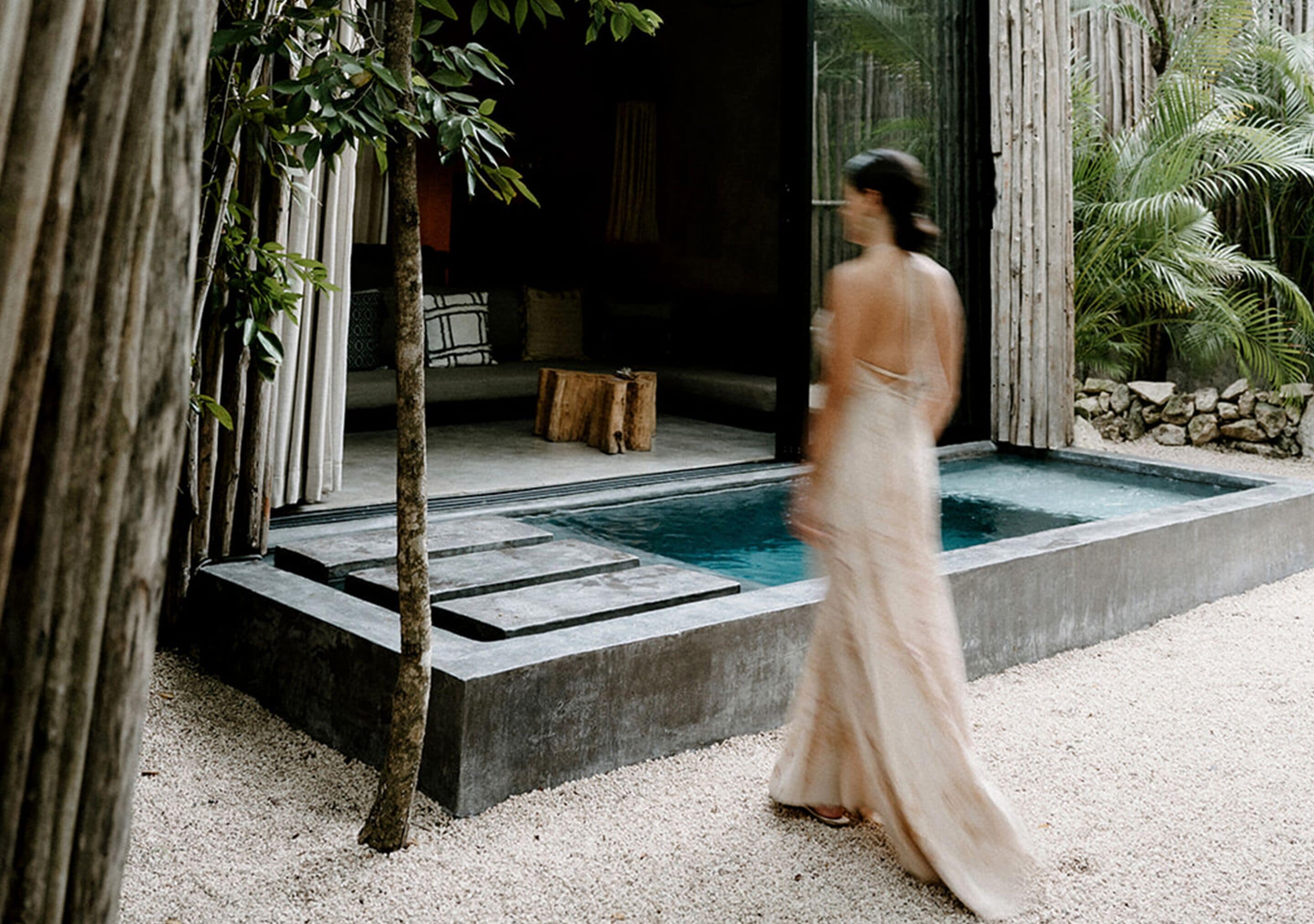 Une femme vêtue d’une longue robe ample se dirige vers un espace piscine au design moderne, entouré d’éléments naturels et bordé d’un mur en pierre.