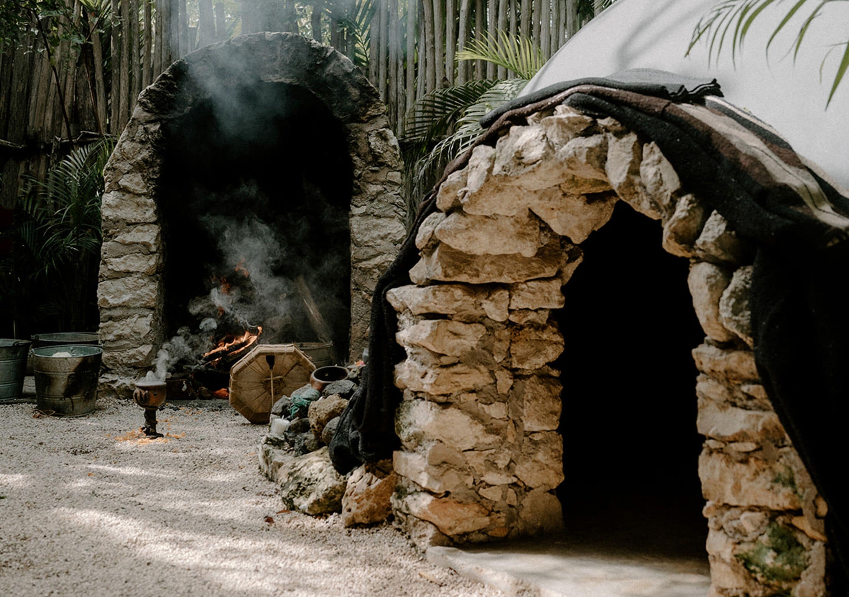 A stone oven with smoke, surrounded by a rustic setting with a bamboo fence and potted plants.