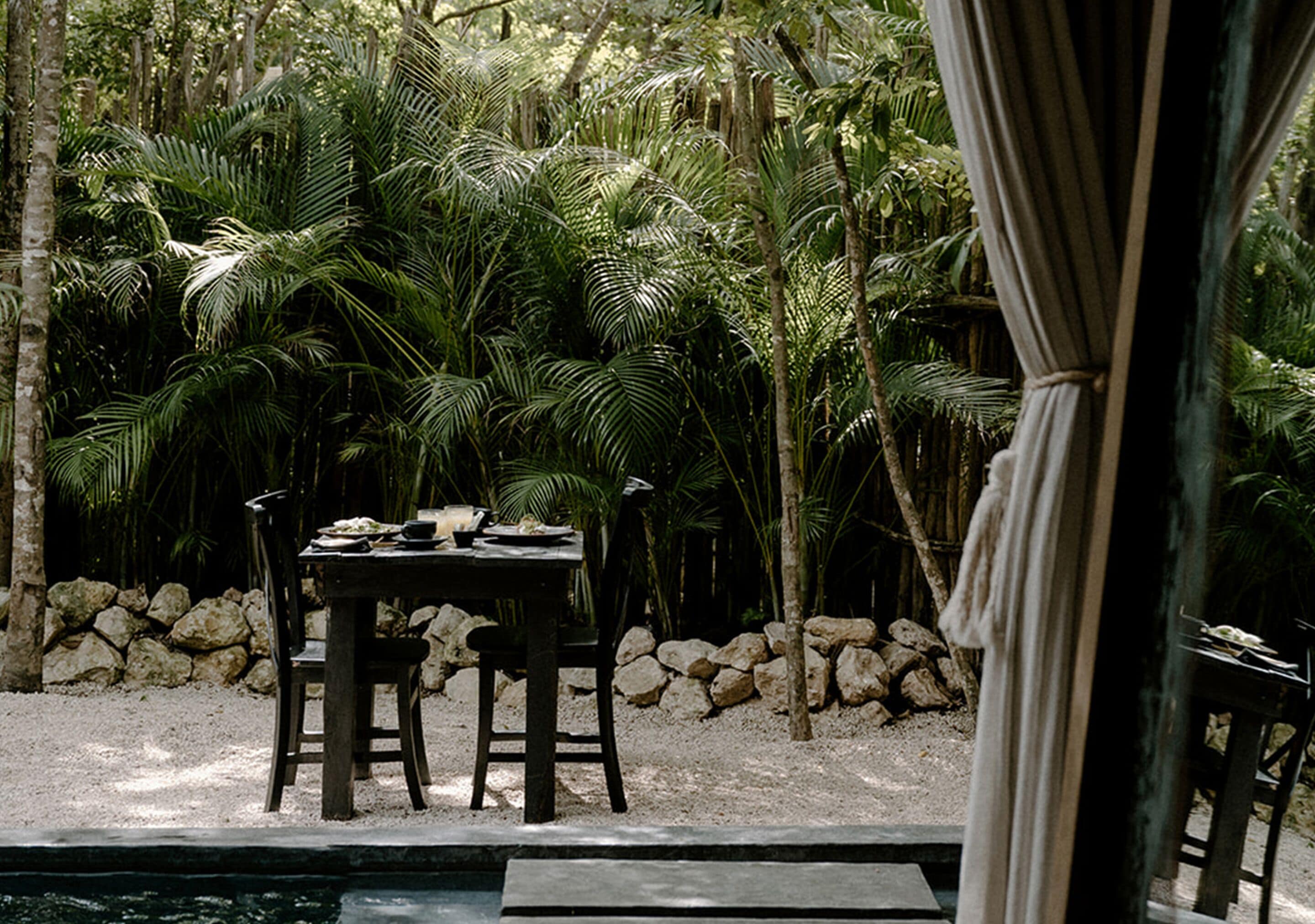 An outdoor dining area with a table and chairs, surrounded by palm trees and a stone wall.