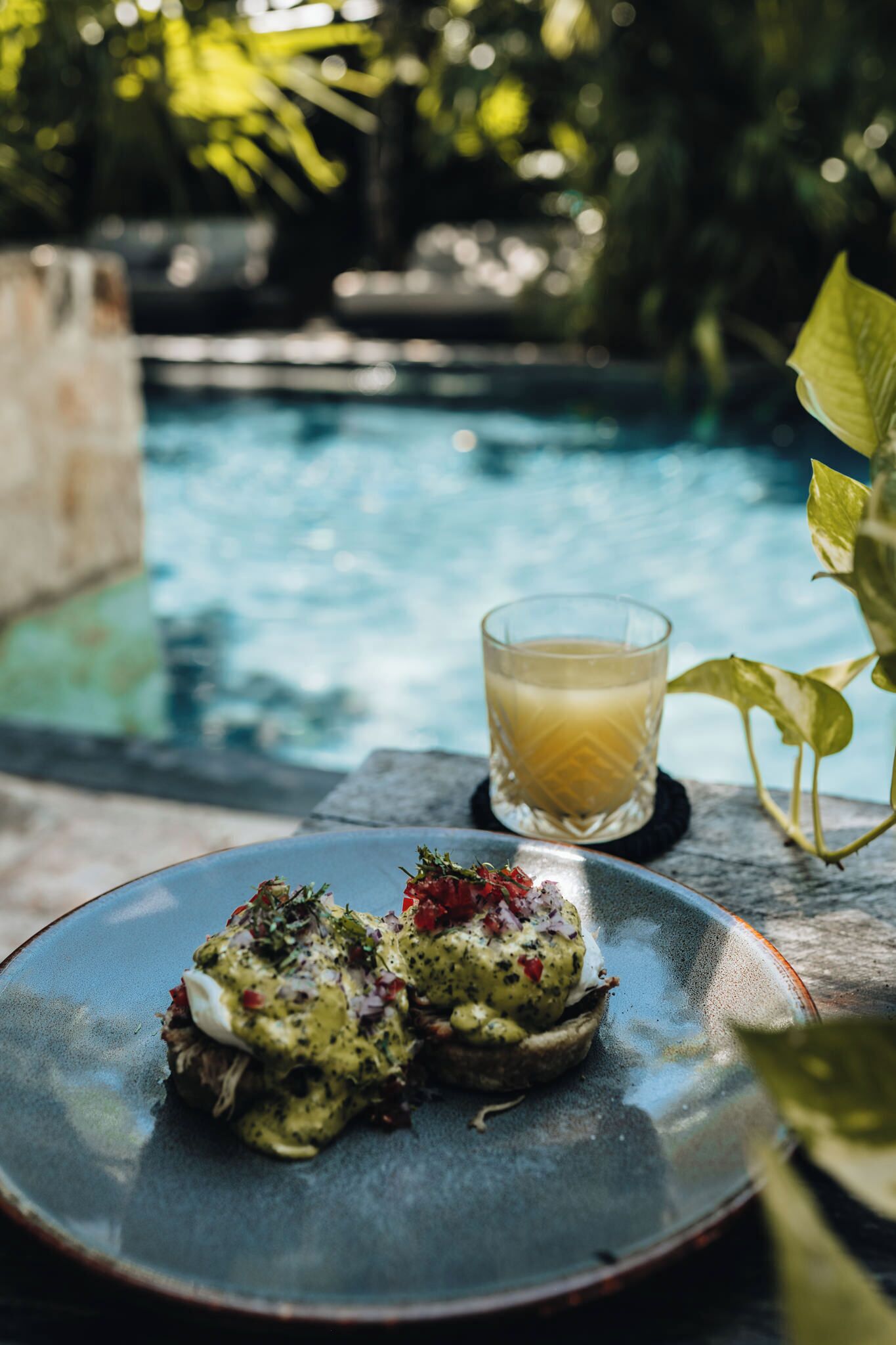 A dining table with a plate of food and a glass of drink by a pool.