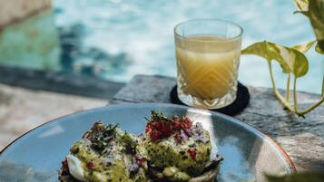 A dining table with a plate of food and a glass of drink by a pool.