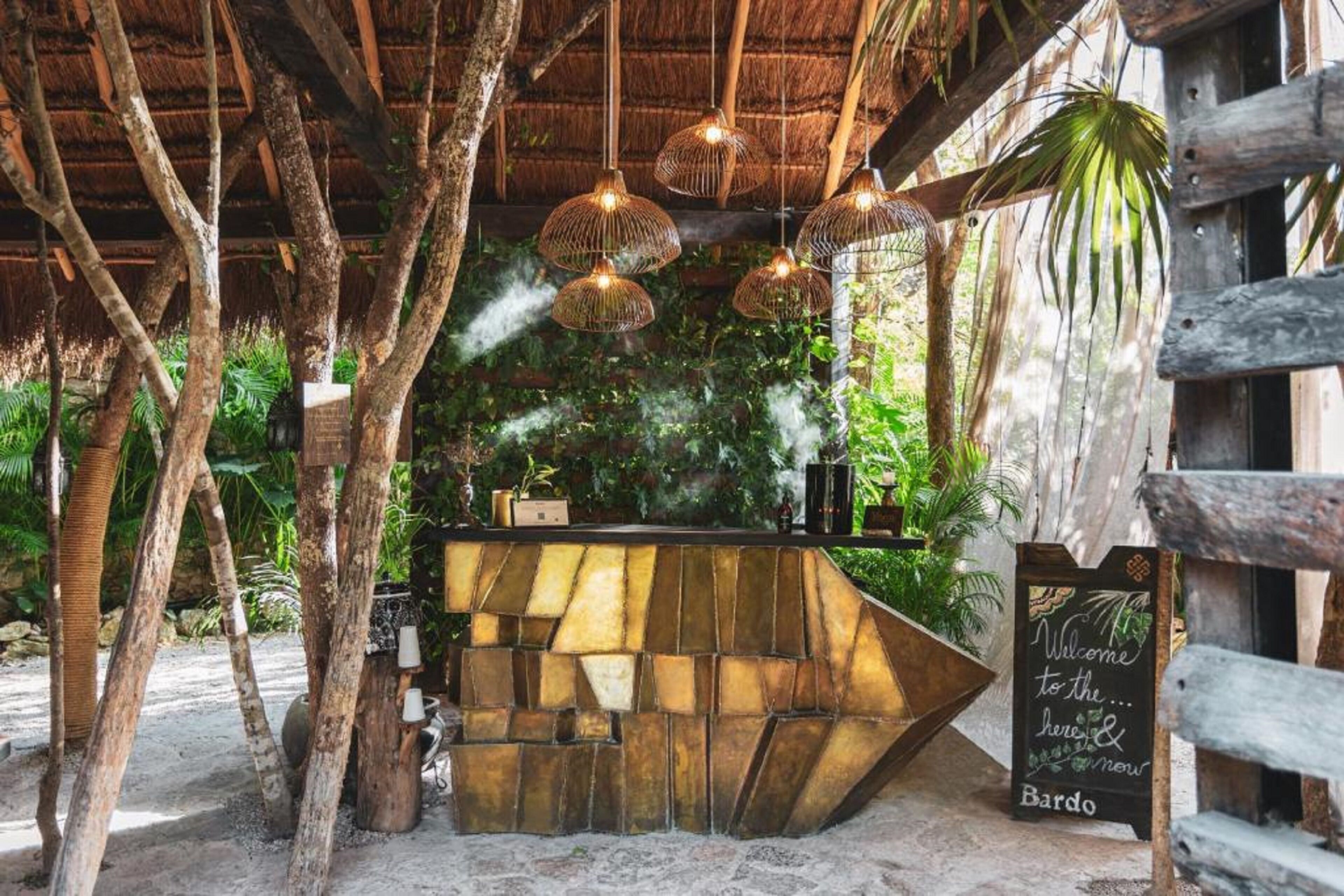 A rustic outdoor bar area with a wooden counter, hanging wicker lamps, and a chalkboard sign welcoming guests to Bardo.