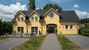 A two-story yellow house with a tiled roof, a front entrance, and a paved driveway. - Hotel Eifellust (Schalkenmehren)