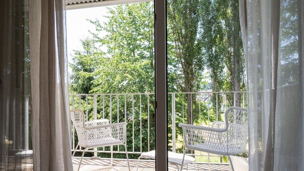 A balcony with white wicker chairs and a table, overlooking a garden with trees and a wooden deck. - Hotel Eifellust (Schalkenmehren)